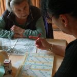 A senior woman and her caregiver work together on a crossword puzzle in a cozy kitchen.