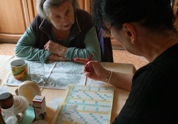 A senior woman and her caregiver work together on a crossword puzzle in a cozy kitchen.