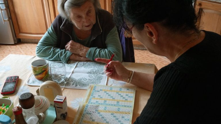 A senior woman and her caregiver work together on a crossword puzzle in a cozy kitchen.