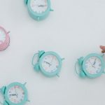 A hand reaching for pastel alarm clocks on a plain white background. Minimalist style.