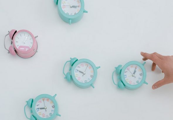 A hand reaching for pastel alarm clocks on a plain white background. Minimalist style.